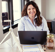 A smiling woman with long brown hair sits at a desk using a black laptop, with a smartphone on a small white tripod to her left and a jar of granola and a bottle of Vielle Serum on the desk to her right.